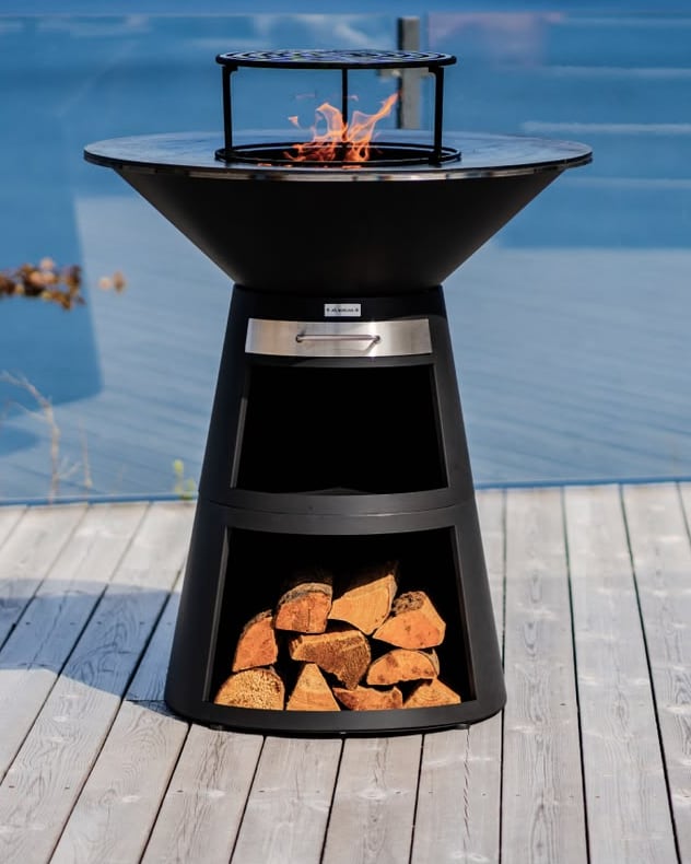 Un brasero allumé, sa plancha et sa réserve de bûches, sur une terrasse en bois.