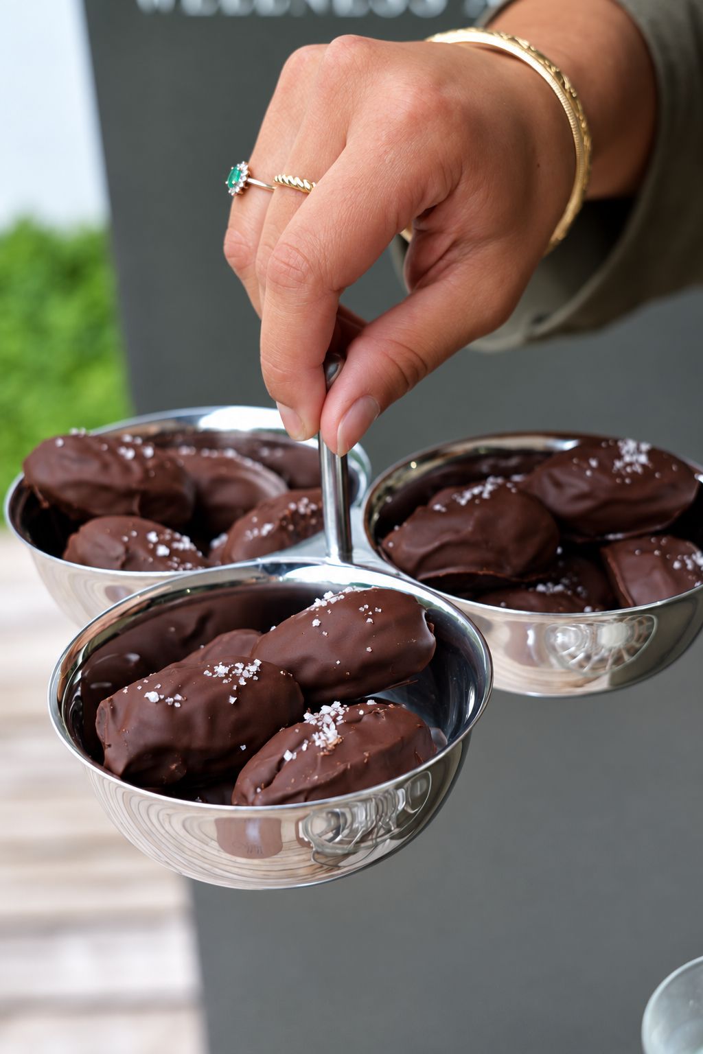 Une main présente trois coupelles argentées de dattes enrobées de chocolat et de fleur de sel.