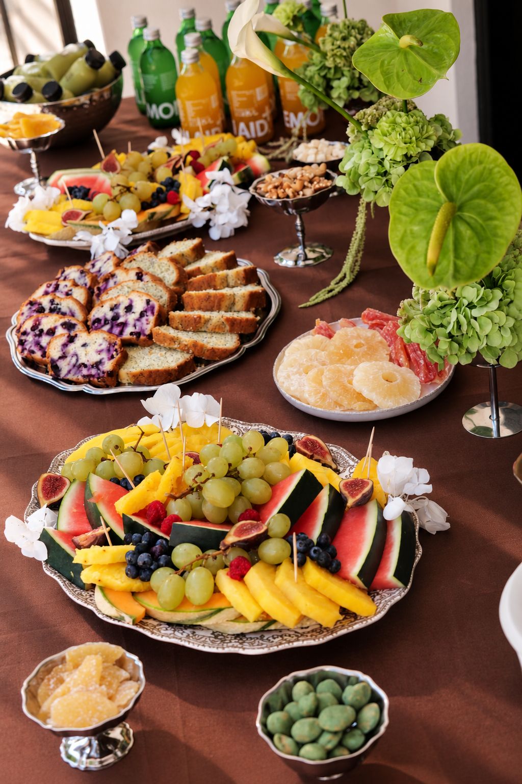Table de brunch nappée de brun : plateaux de fruits frais, cakes et fleurs vertes.