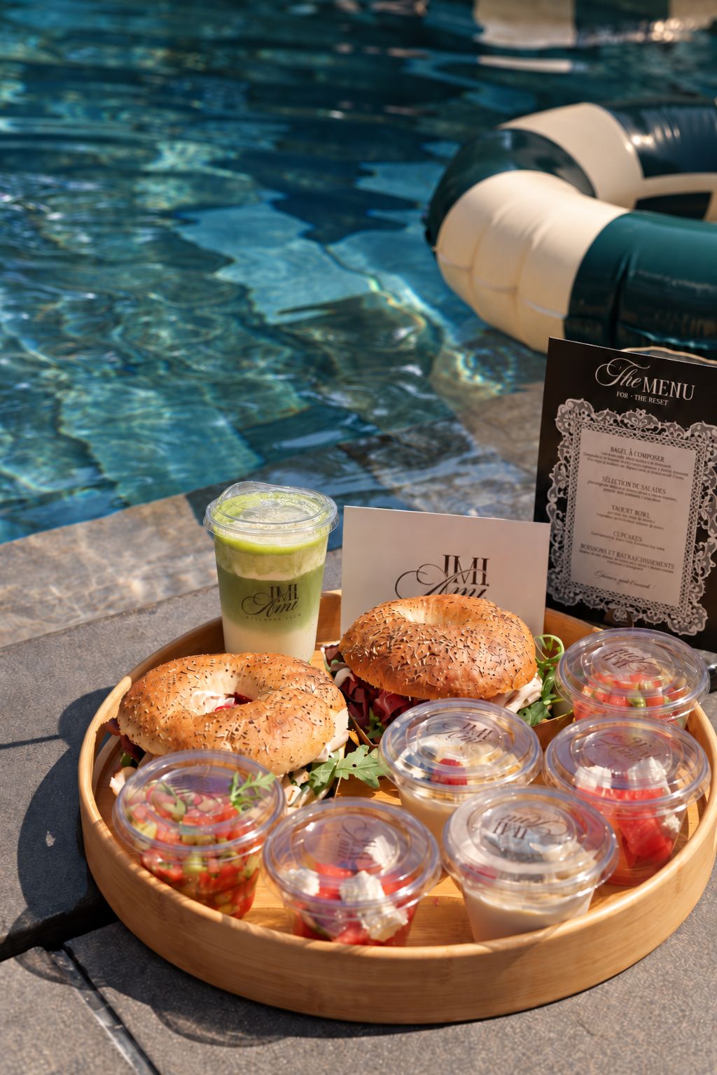 Plateau de brunch au bord de la piscine : bagels, salades en pots et matcha latte au logo de la marque.
