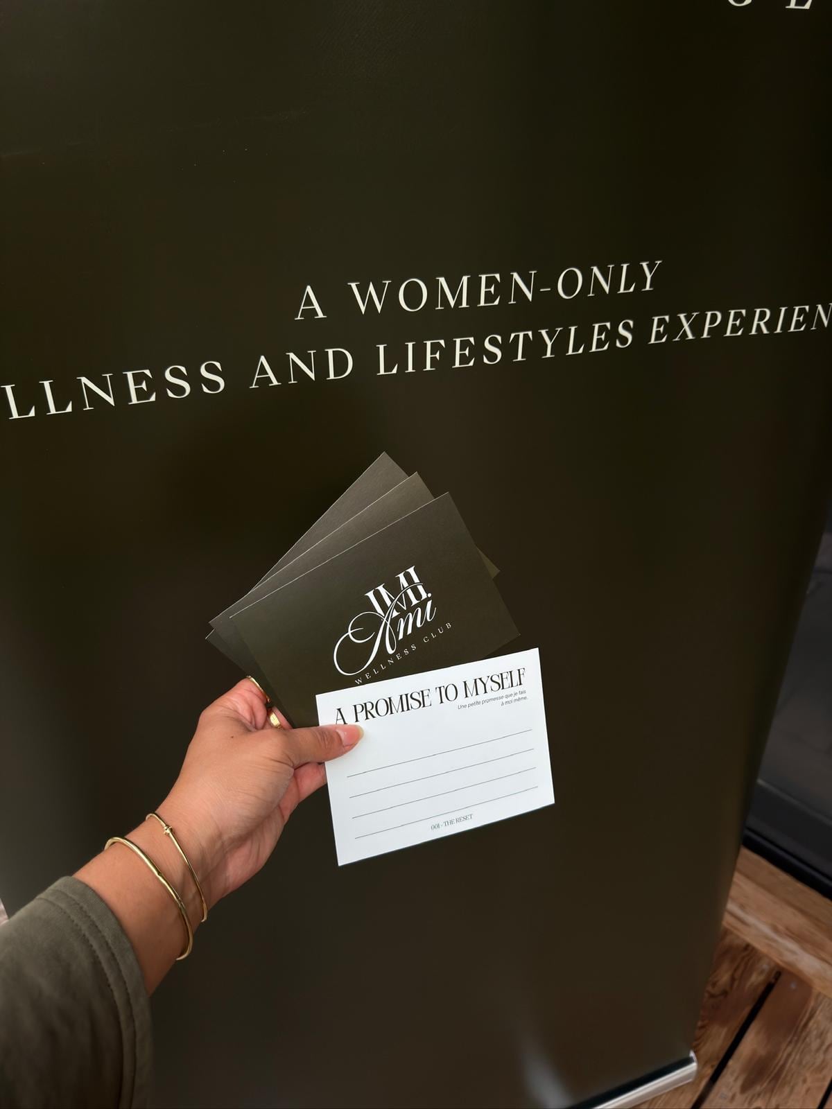 Une main tient les cartes « A promise to myself » de l'édition 001, devant le kakémono « a women-only wellness and lifestyles experience ».