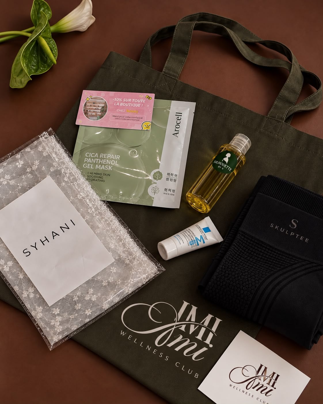 Le tote bag olive au logo IMI Ami, à plat, entouré de son contenu : pochette Syhani, masque, huile de soin et textile de sport.