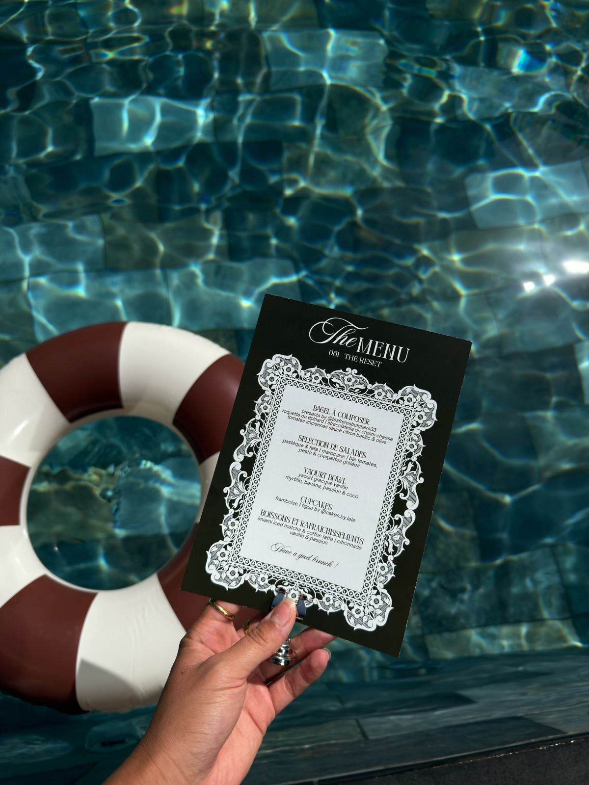 Une main tient le carton « The Menu — 001 The Reset » au-dessus de l'eau de la piscine.